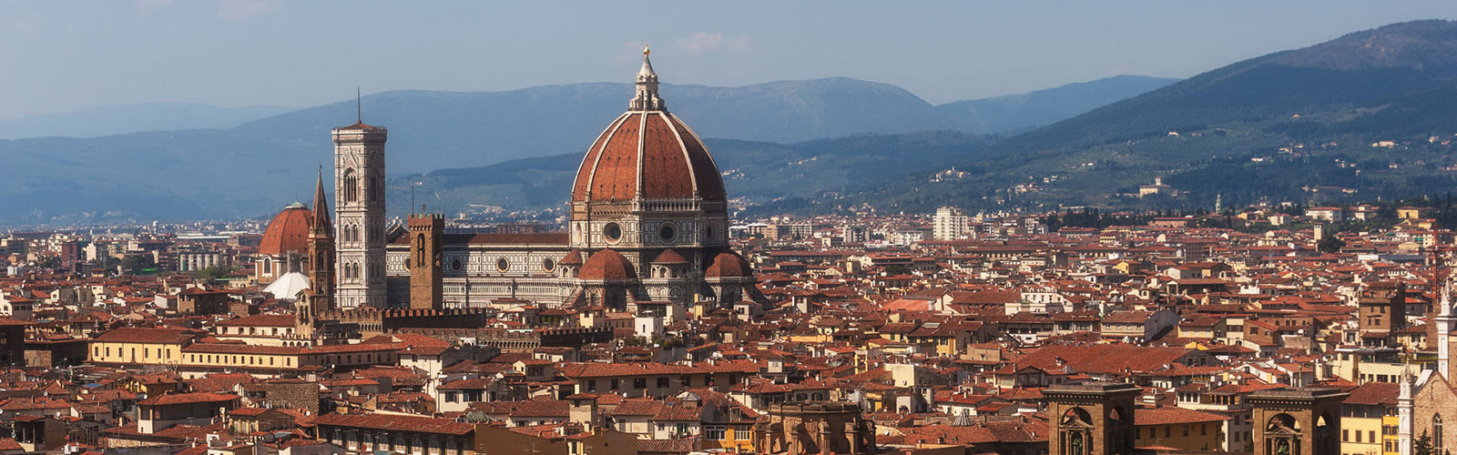 A city full of buildings with reddish-brown roofs, with one large stone building in the middle of the city, with a circular domed roof. There are green mountains visible in the background. 